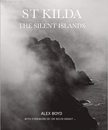 The book cover of St Kilda: The Silent Islands by Alex Boyd is black and white. It features a photograph depicting a rugged coastline with high cliffs, partially obscured by fog or mist. The landscape shown includes visible rock formations and the presence of dense atmospheric conditions, which limit visibility in parts of the scene. The title and authors name are displayed on the cover, though placement and typography details may vary by edition.