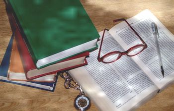 A stack of colorful books, an open book, red glasses, a pen, and a pocket watch are arranged on wood. Sunlight casts dappled shadows over the scene. This setup advertises Genealogy Research (2 Hours) for £60.
