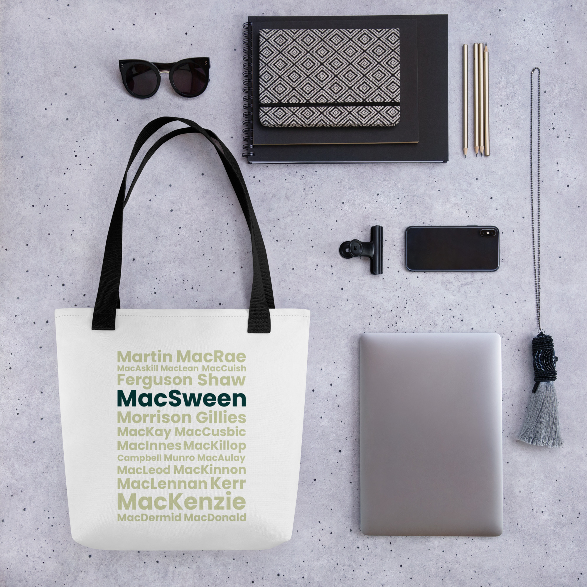 The A Tote bag - MacSween features Scottish surnames printed in green and black. The bag is positioned on a speckled surface. Surrounding the tote are a laptop, a notebook, a pen, a phone, a camera lens, a tasseled keychain, sunglasses, and a patterned pouch.