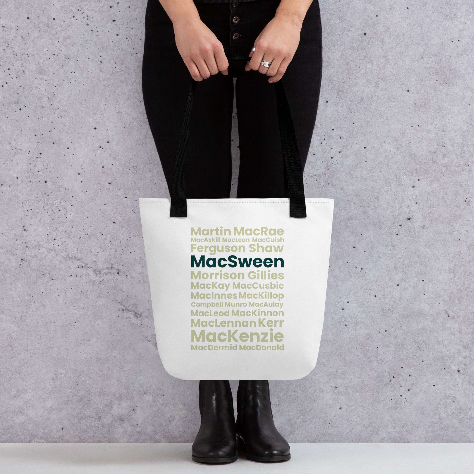 A person holds the Tote bag - MacSween by its black straps. The tote displays Scottish last names in green and gray, with MacSween prominently highlighted. The individual is wearing black pants and boots.