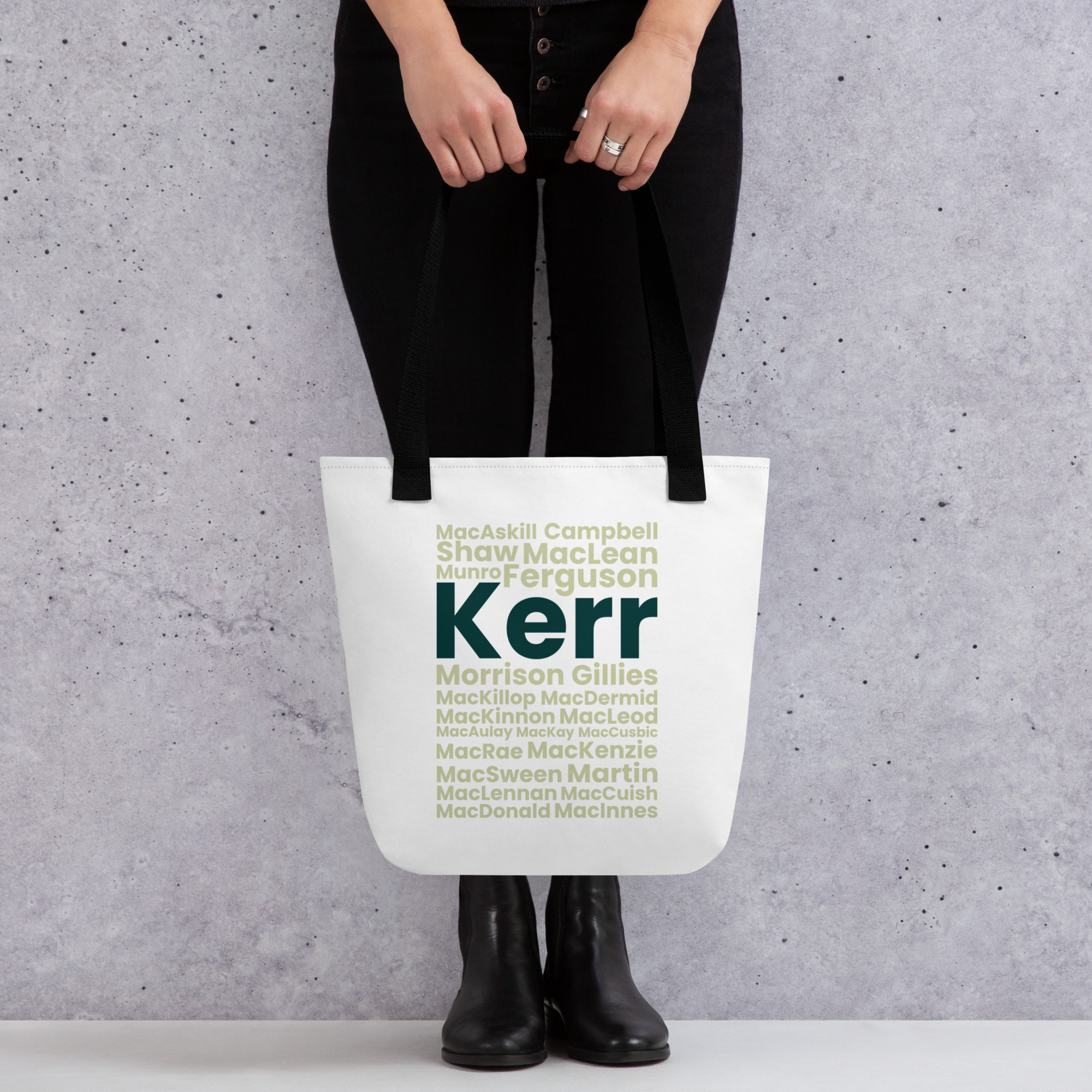 A person in black pants and boots holds the Tote bag - Kerr. The tote features a white surface printed with green and teal surnames, including Kerr displayed in bold letters. The background behind the person is gray and speckled.