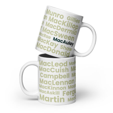 Two white mugs are shown, each with a light green Scottish surname printed on them. The mugs are stacked, and the top mug features MacAulay in bold black text. The Mug - MacAulay is described as a distinctive gift item for those interested in collecting or using mugs.
