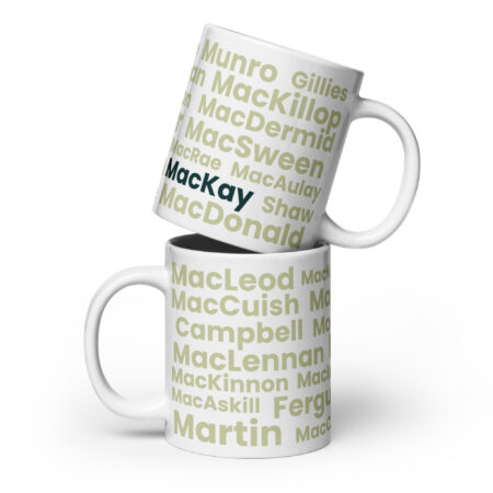 Two white mugs are stacked, each displaying the Mug - MacKay design. This design features Scottish surnames—MacLeod, Campbell, Munro, and Martin—in green and gray lettering. The name MacKay is presented prominently in bold text, creating a striking visual emphasis on that surname.