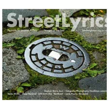 A metal manhole cover with geometric patterns is set among grass and cobblestones. It displays credits for "Street Lyrics.
