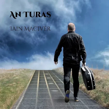 A man in black walks away on a road carrying a guitar case under a cloudy sky. "AN TURAS" and "IAIN MACTVER" appear above him.