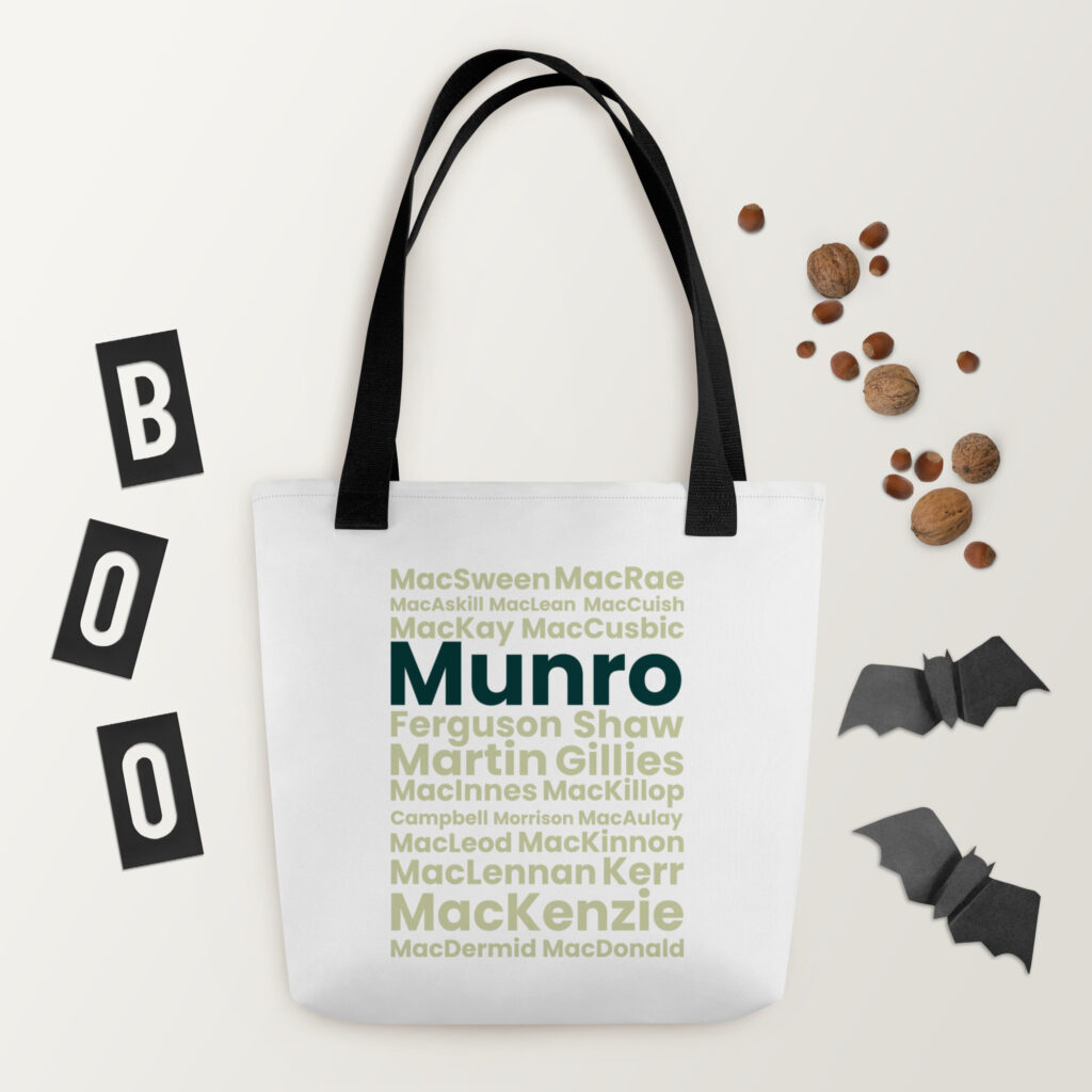 The Tote bag - Munro is a white tote with black handles. It features Scottish surnames, such as Munro and MacKenzie, printed in green text. In the described setting, the tote is styled with black paper bats, nuts, and “BOO” letters on or around it. The arrangement stands out against a light background.