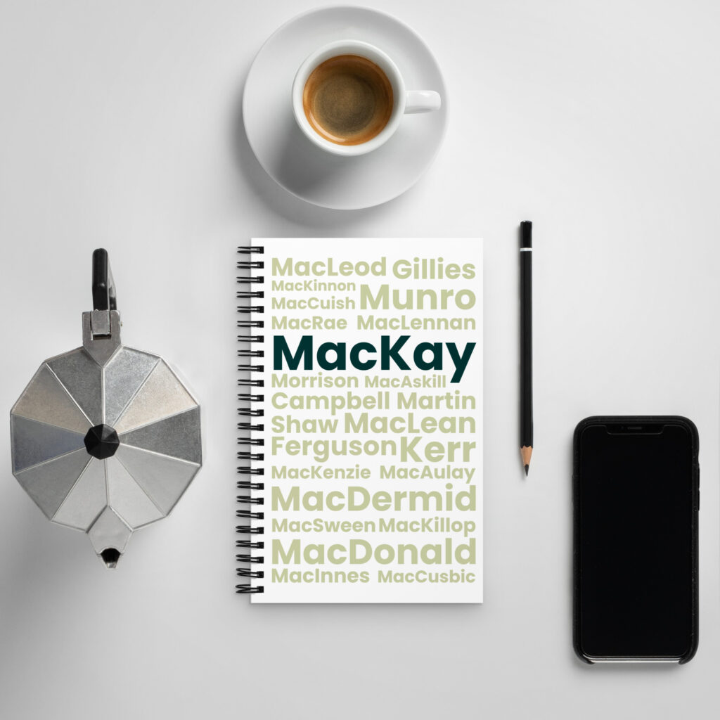 The flat lay image features a notepad with the surname MacKay prominently displayed. The arrangement also includes a cup of coffee, a moka pot, a black pen, and a smartphone. All items are positioned on a white table.