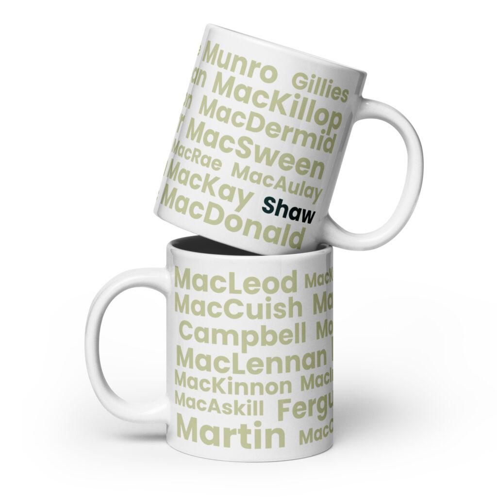 Two white Mug - Shaw mugs are stacked. Each mug is printed with multiple Scottish surnames in light green text. On the top mug, the surname Shaw is presented in bold black lettering, while the other surnames remain in light green.