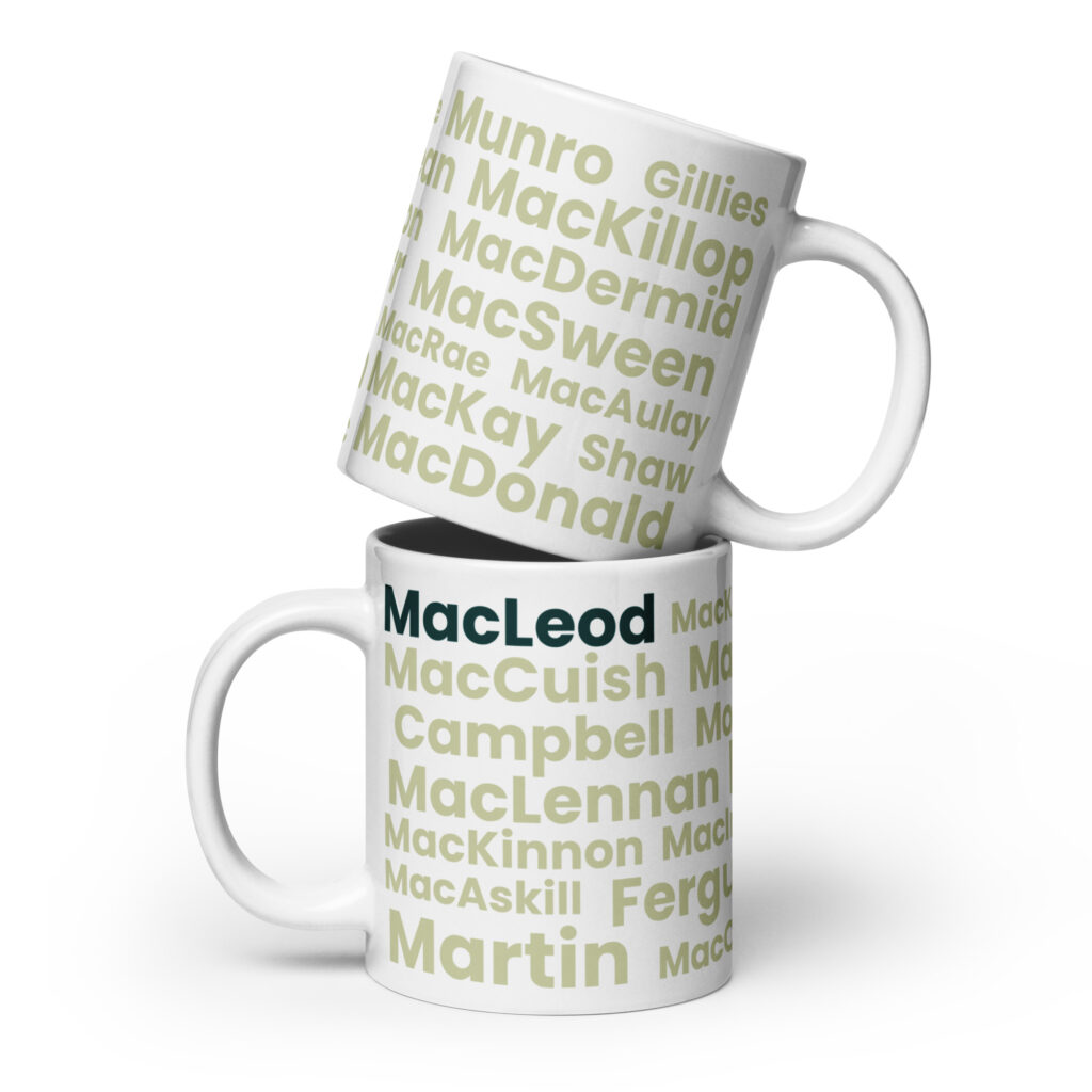 Two white mugs labeled MacLeod are stacked together. The bottom mug is upright, while the top mug rests at an angle with its rim on the lower mug. Each mug displays a series of Scottish surnames, with MacLeod prominently highlighted in bold dark green lettering.