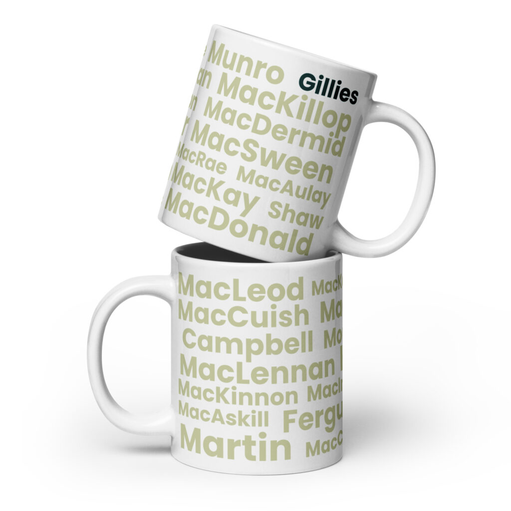 The Mug - Gillies features a design displaying a list of Scottish surnames, including MacDonald, MacLeod, and Martin. The surnames are printed in light green text on a white background. The mug labeled Gillies is distinguished by darker green text, highlighted more prominently than the others. In the image or arrangement described, one mug is positioned on top of another.