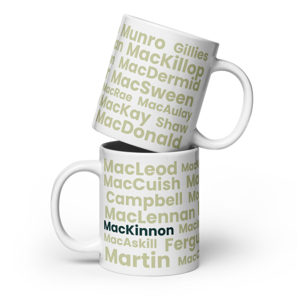 Two white ceramic mugs are shown. Both feature light green text with Scottish surnames. The lower mug is labeled “MacKinnon” in bold black letters and is positioned on top of the stack. The design highlights Scottish heritage, specifically associated with the MacKinnon surname.